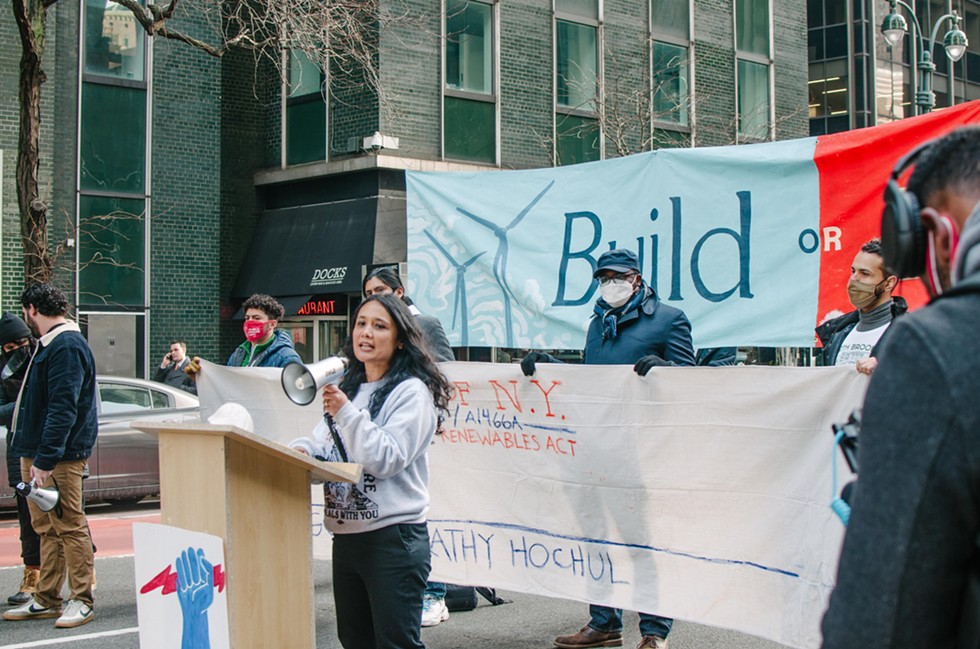 A woman in a sweatshirt and jeans speaking into a megaphone at a street rally, in front of a banner reading Build or Burn.