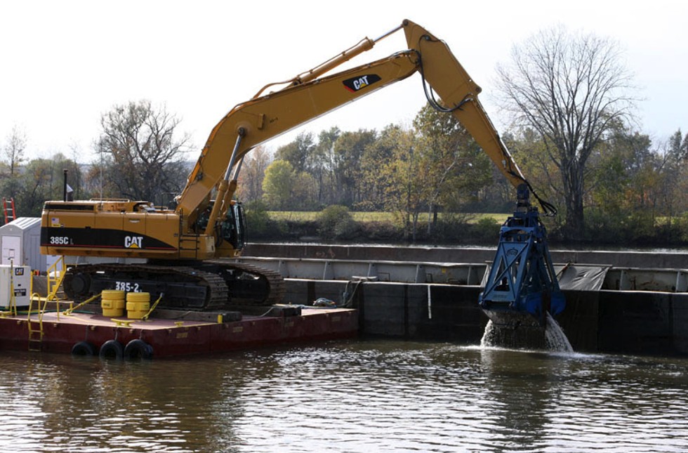 Workers use excavators with environmental clamshell buckets mounted on flat, anchored platforms to dredge the river. The PCB-contaminated sediment is emptied onto 35-foot-wide, 195-foot-long floating barges.