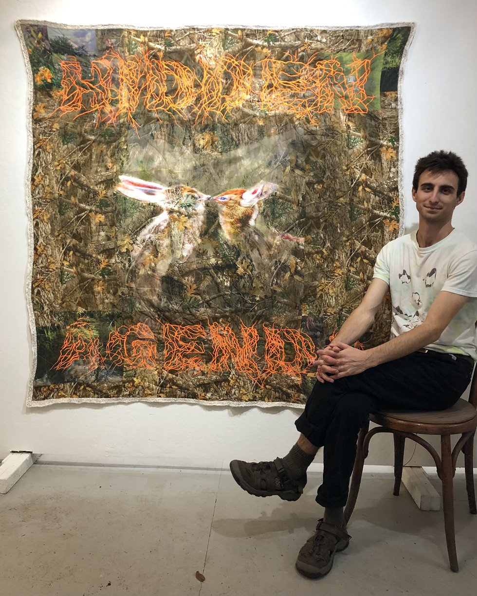 The artist, John DeSousa, sits with his embroidered quilt, titled, "HIDDEN AGENDA"