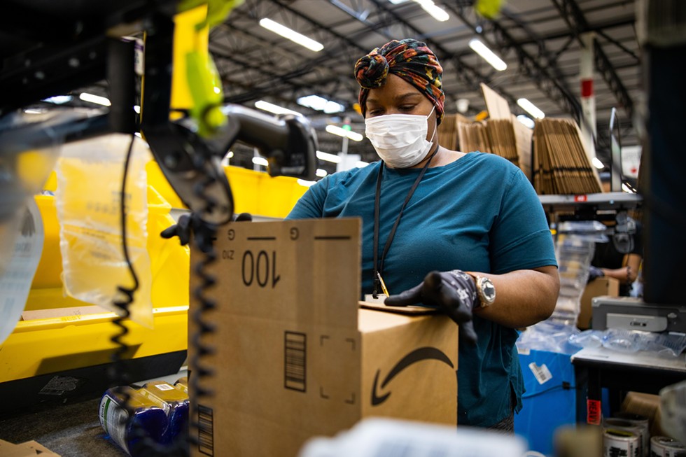 A woman wearing PPE boxing up an Amazon package on a conveyor belt.